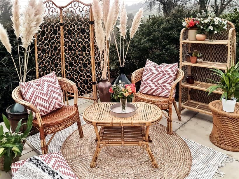 Rattan furniture on a jute rugs in the garden surrounded by a rattan screen, a rattan bookcase and lots of potted plants and pampas grass