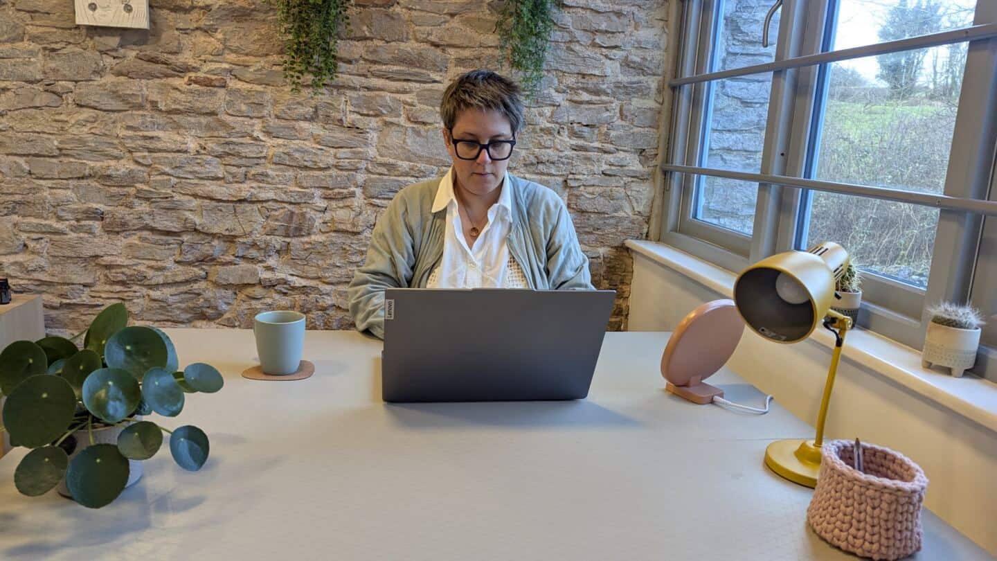 A middle aged woman sits at a desk by the window working on a laptop next to a SAD light