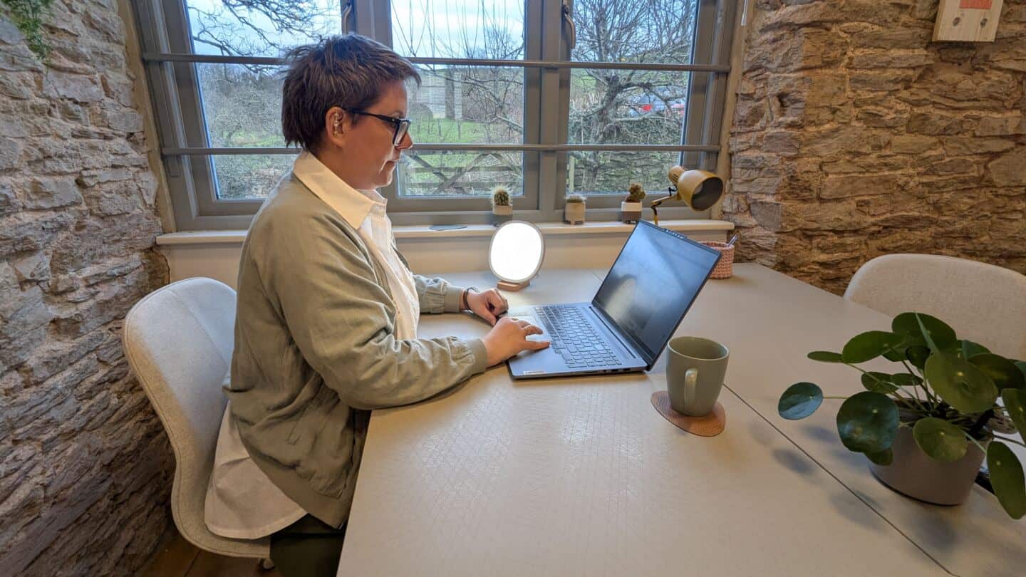 A middle aged woman sits at a desk by the window working on a laptop next to a SAD lamp