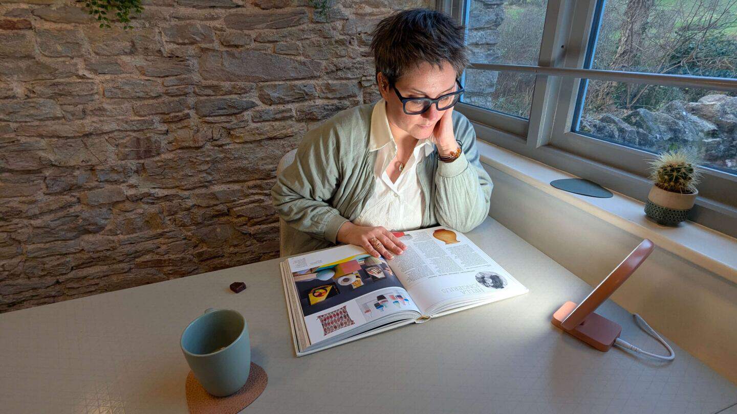 A middle aged woman sits at a desk by the window reading a book next to a SAD light