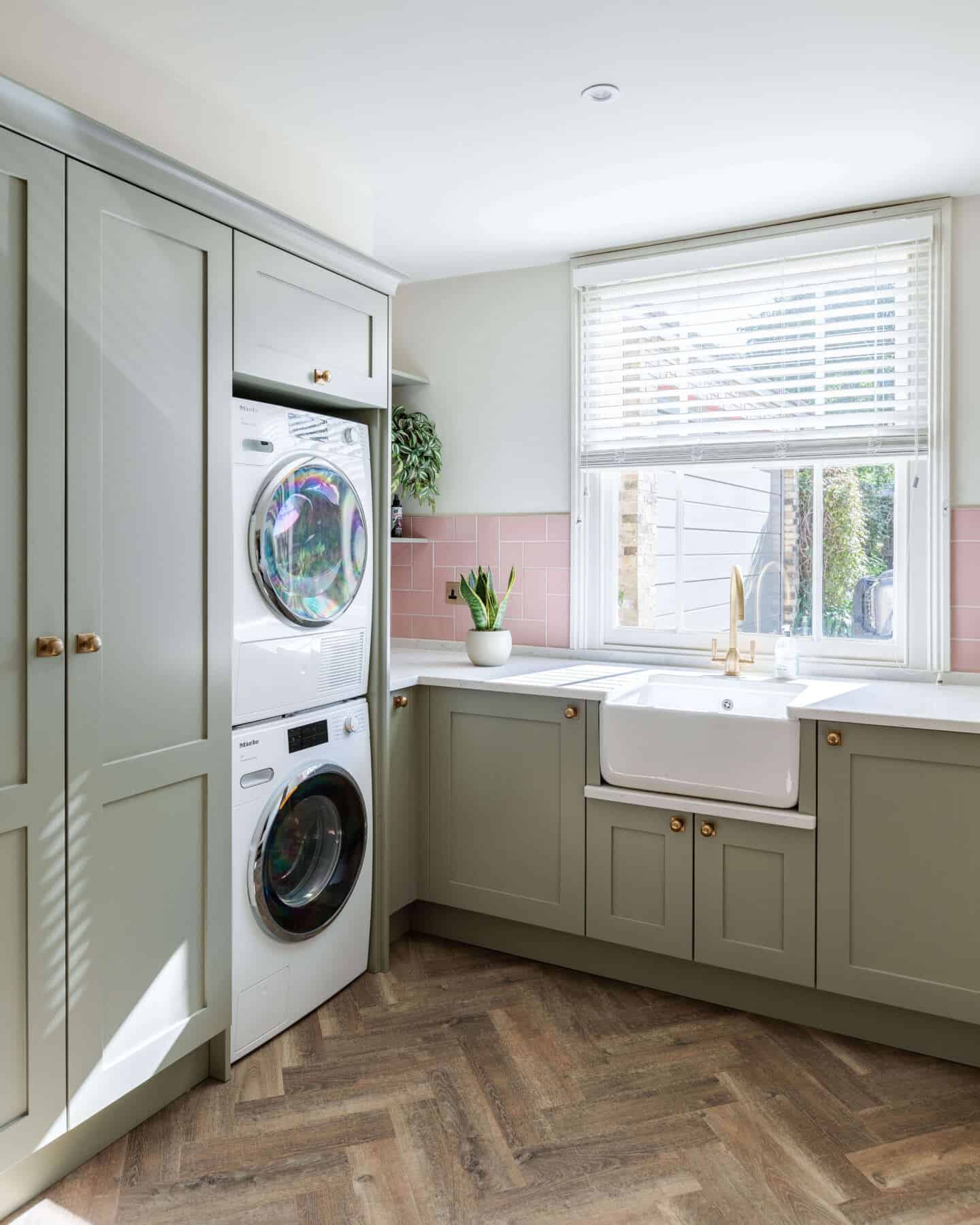 A sage green utility room with wooden herringbone flooring, a large ceramic butler sink and a tumble dryer stacked on top of a washing machine