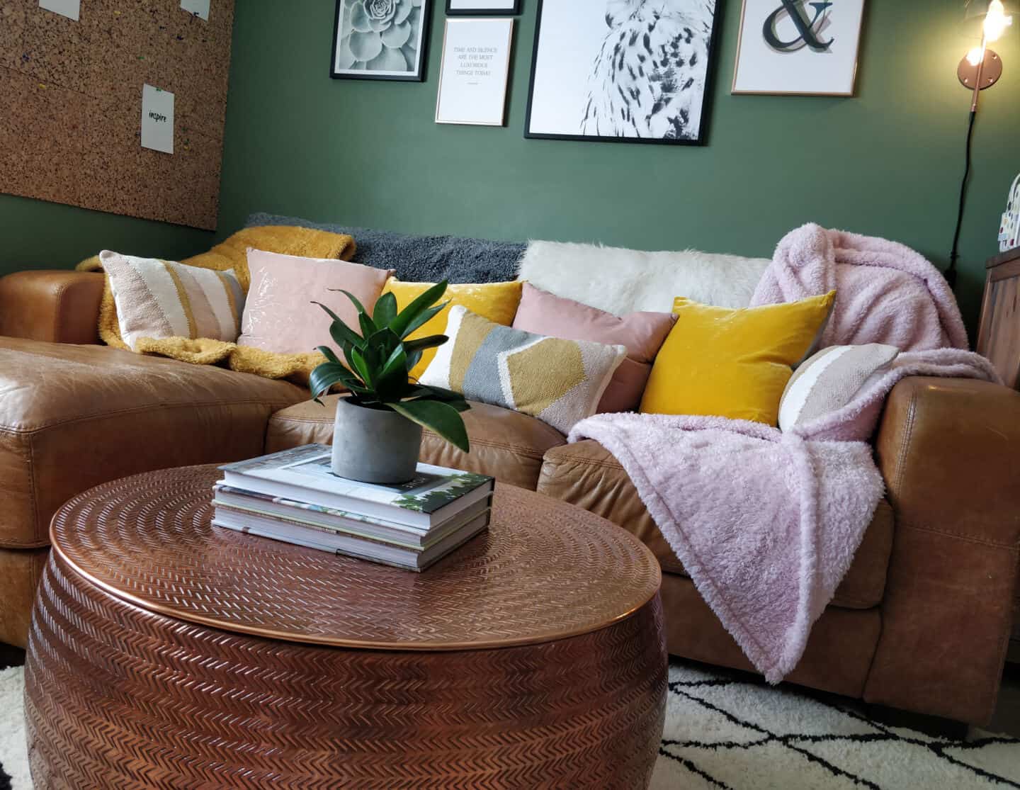 A small living room with dark green walls, a round storage copper coffee table and a brown leather sofa covered in textured cushions and blankets