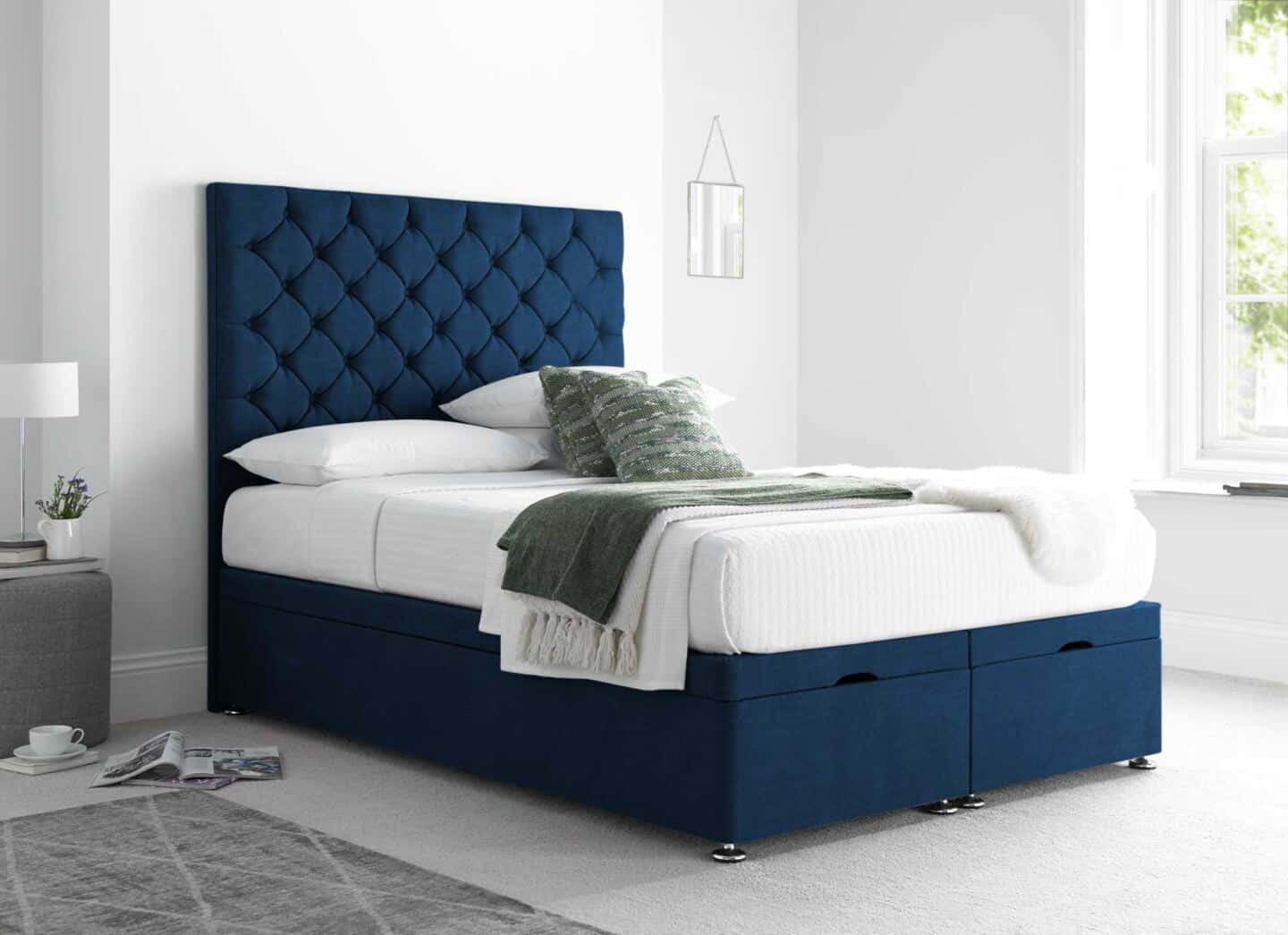 A blue ottoman bed with white bedding and a tall tufted headboard in a large white airy bedroom