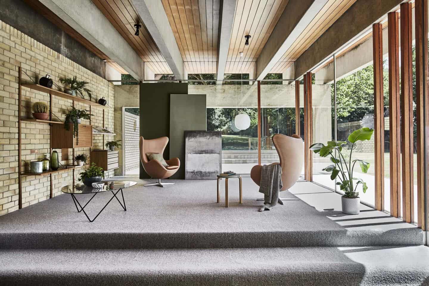 A large open plan mid-century modern living room with silver Berber wool carpet, brick walls, floor to ceiling windows and two Hans Wegner egg chairs