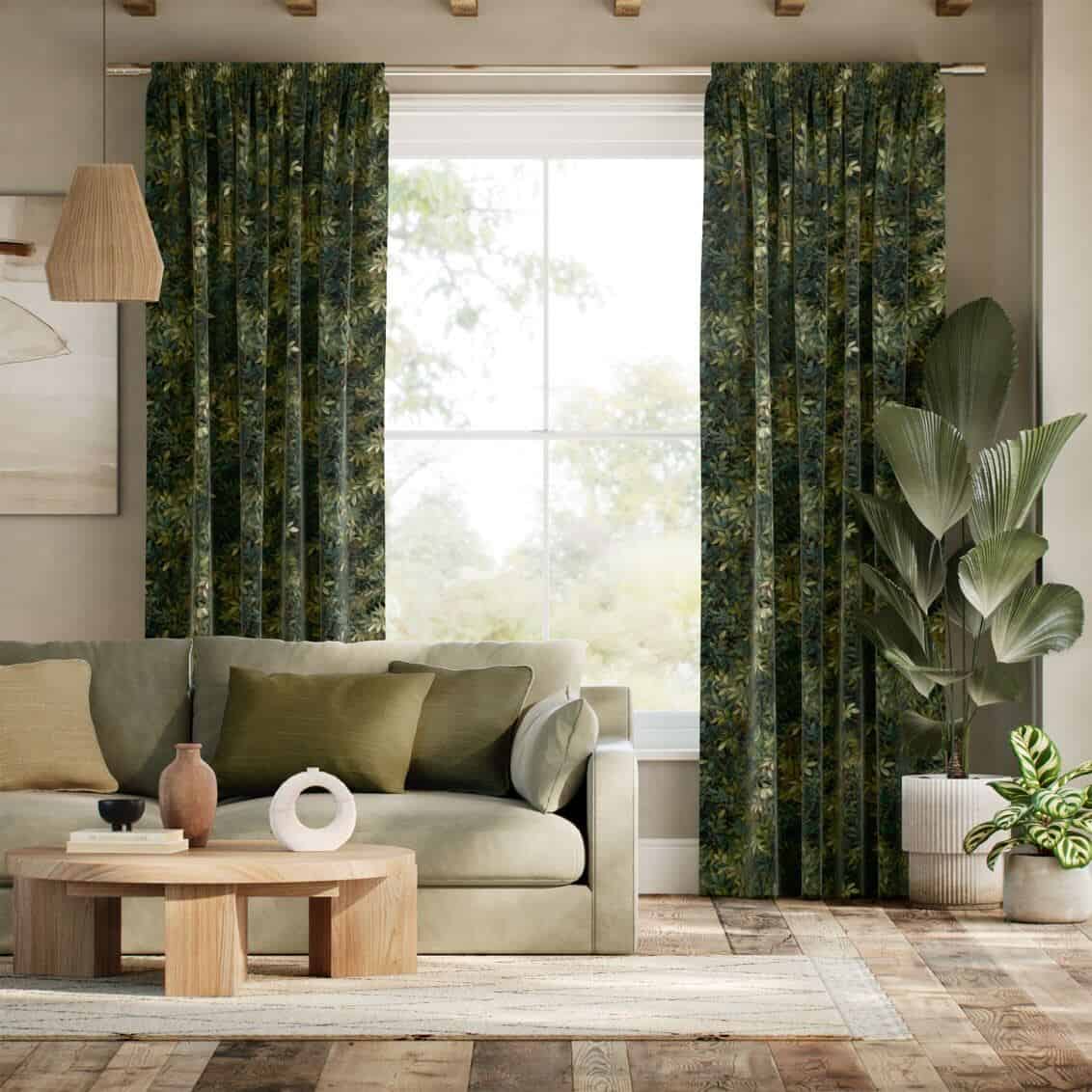 Full length green velvet curtains with leaf print hang in front of a window. A plant and a cream sofa are positioned in front of the window