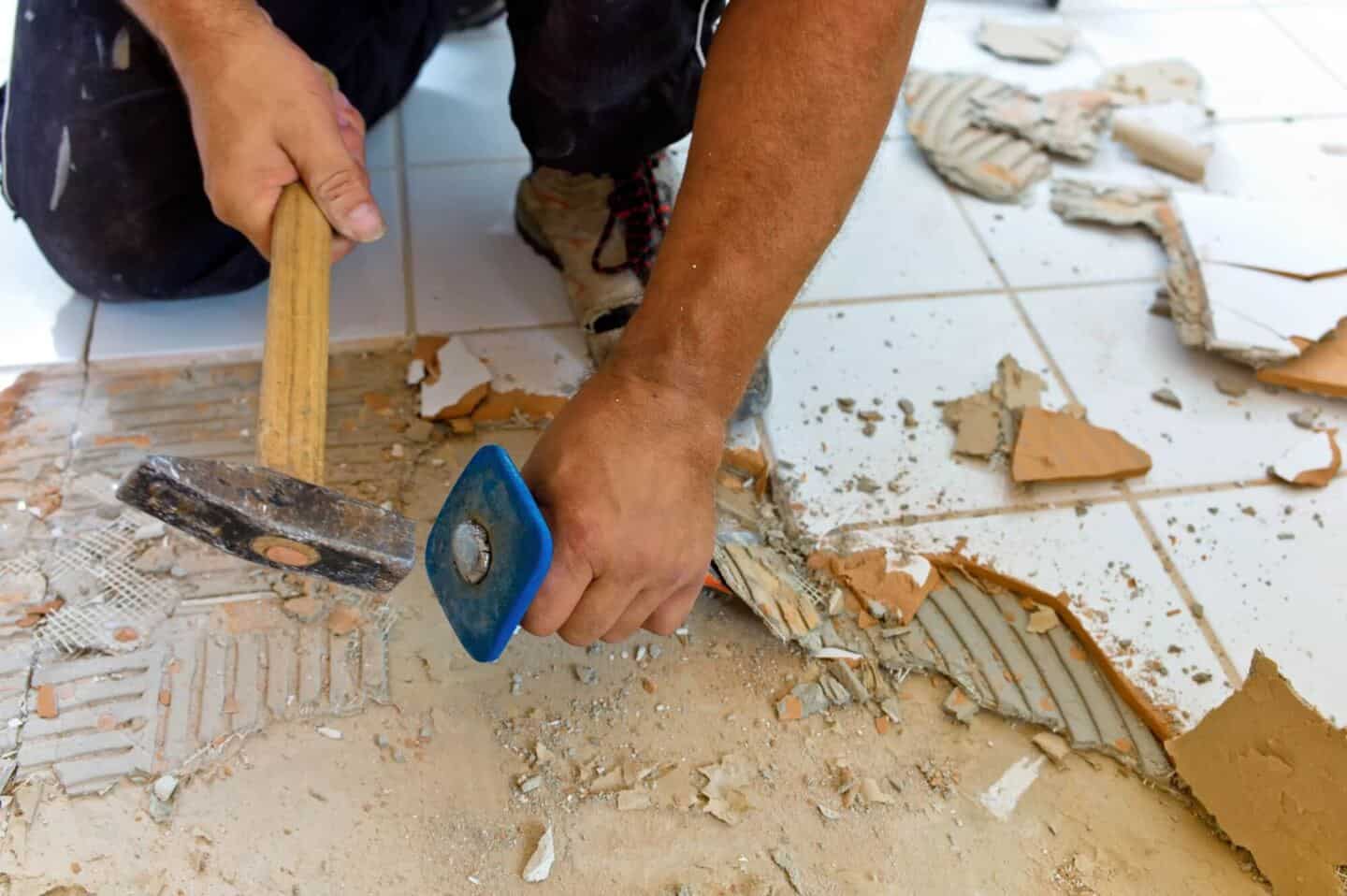 A man on his knees holding a chisel and hammer to scrape ceramic tiles off the floor ready for renovation