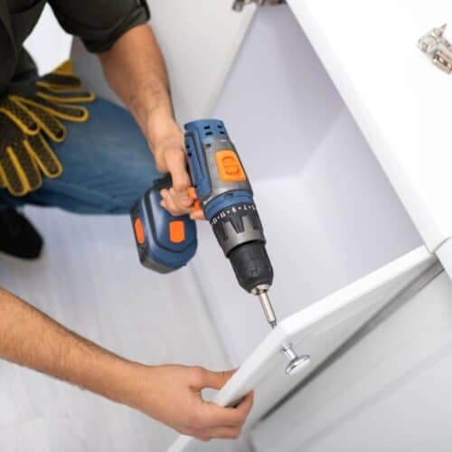 A man crouching down holding a drill and replacing cupboard door knobs