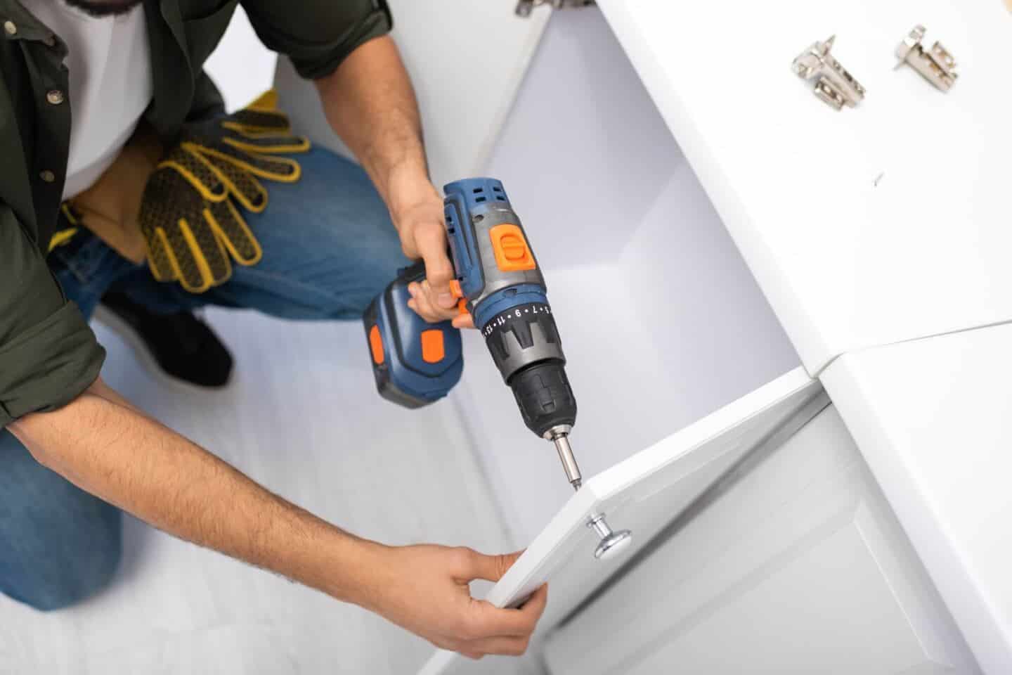 A man crouching down holding a drill and replacing cupboard door knobs