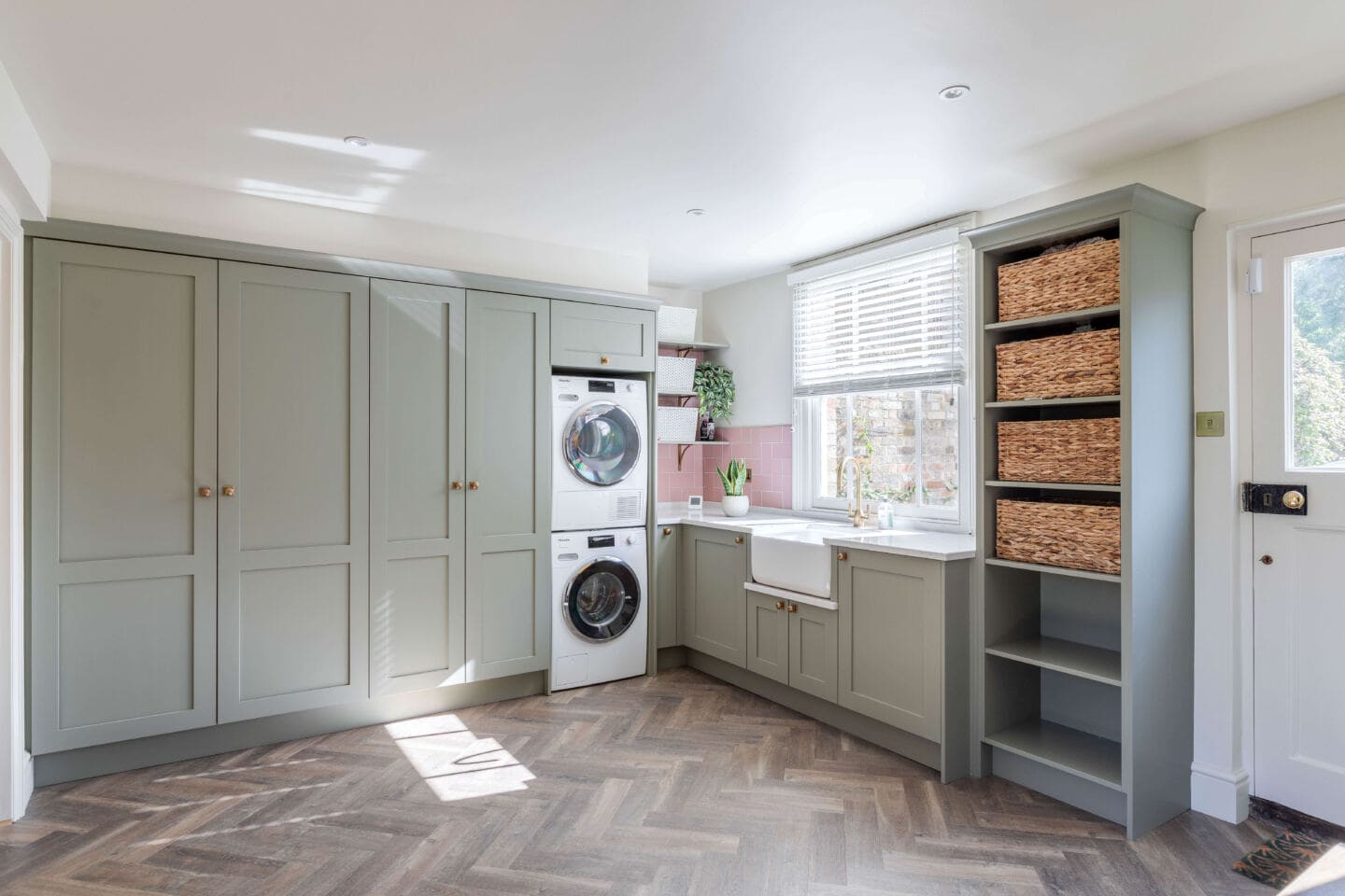 A sage green utility room with wooden herringbone flooring, a large ceramic butler sink and a tumble dryer stacked on top of a washing machine