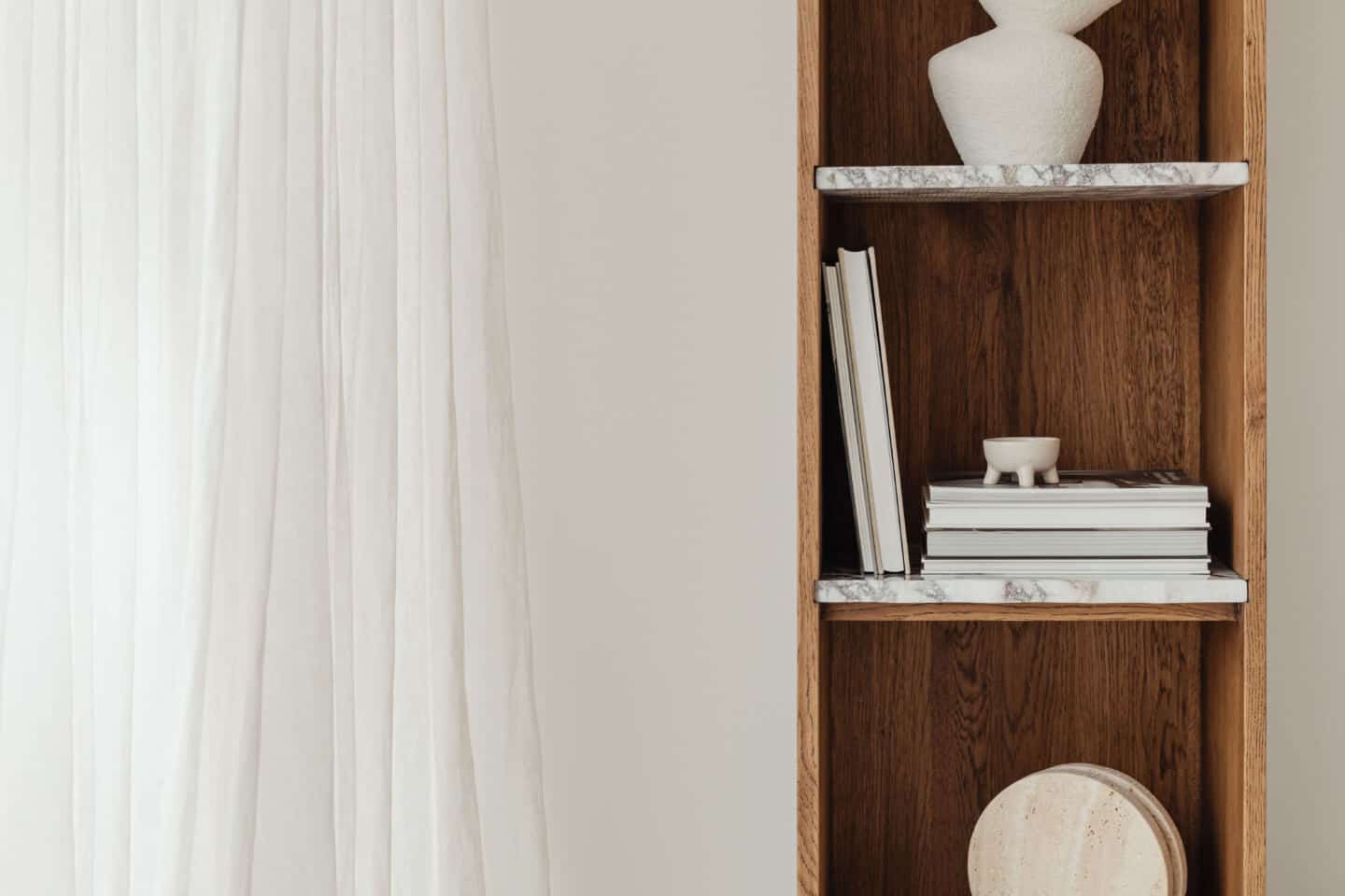 A wooden shelving unit with marble shelves next to a window with white voile curtains