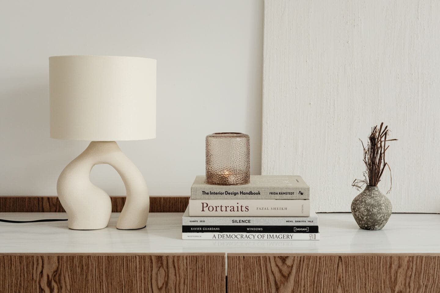 A candle sits on top of a pile of books with an abstract lamp beside and a small vase of dried twigs