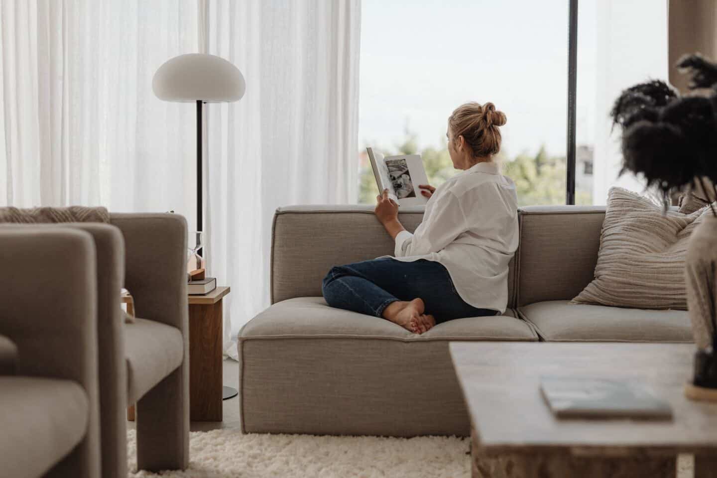Middle aged white woman sat in a calming living space reading a book on a sofa