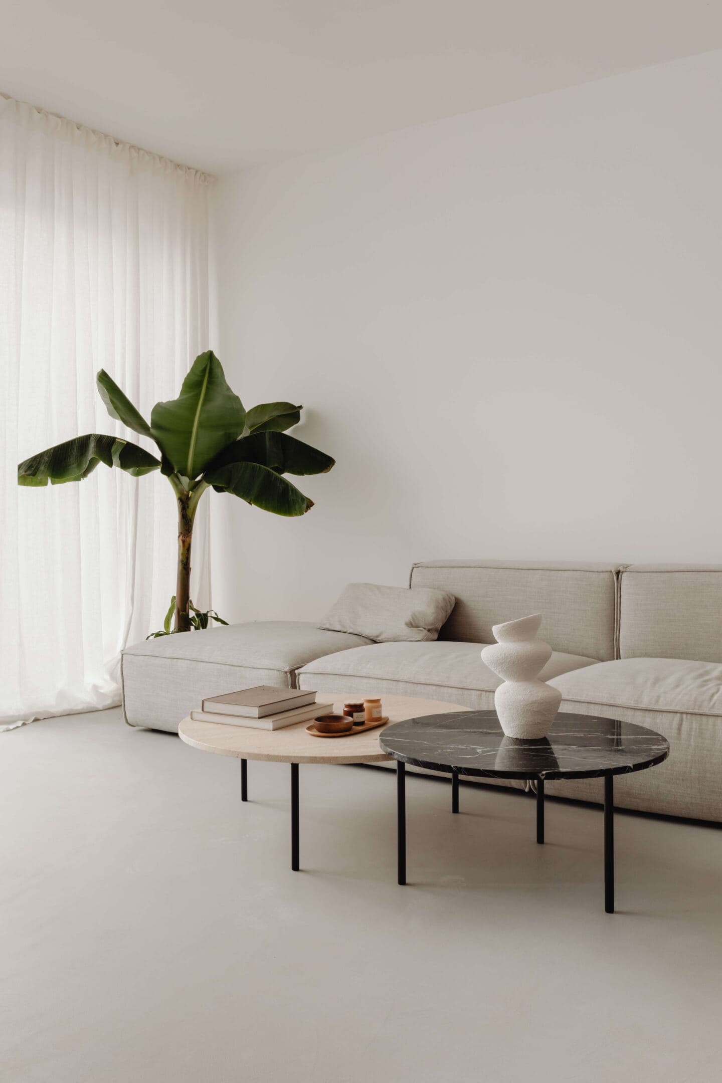 A calming living space with grey boxy grey sofa, two round marble coffee tables and a large banana plant