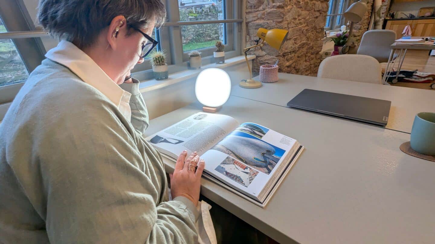 A middle aged woman sits at a desk by the window reading a book next to a SAD light