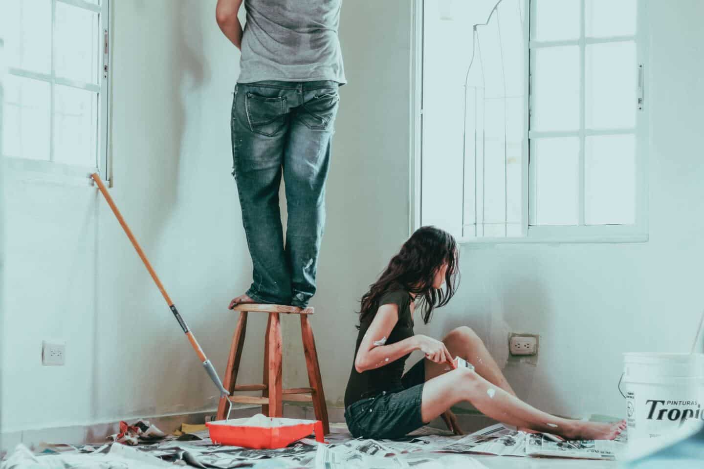 A man stands on a stall and a woman sits on newspaper on the floor. Both are painting the walls with white paint