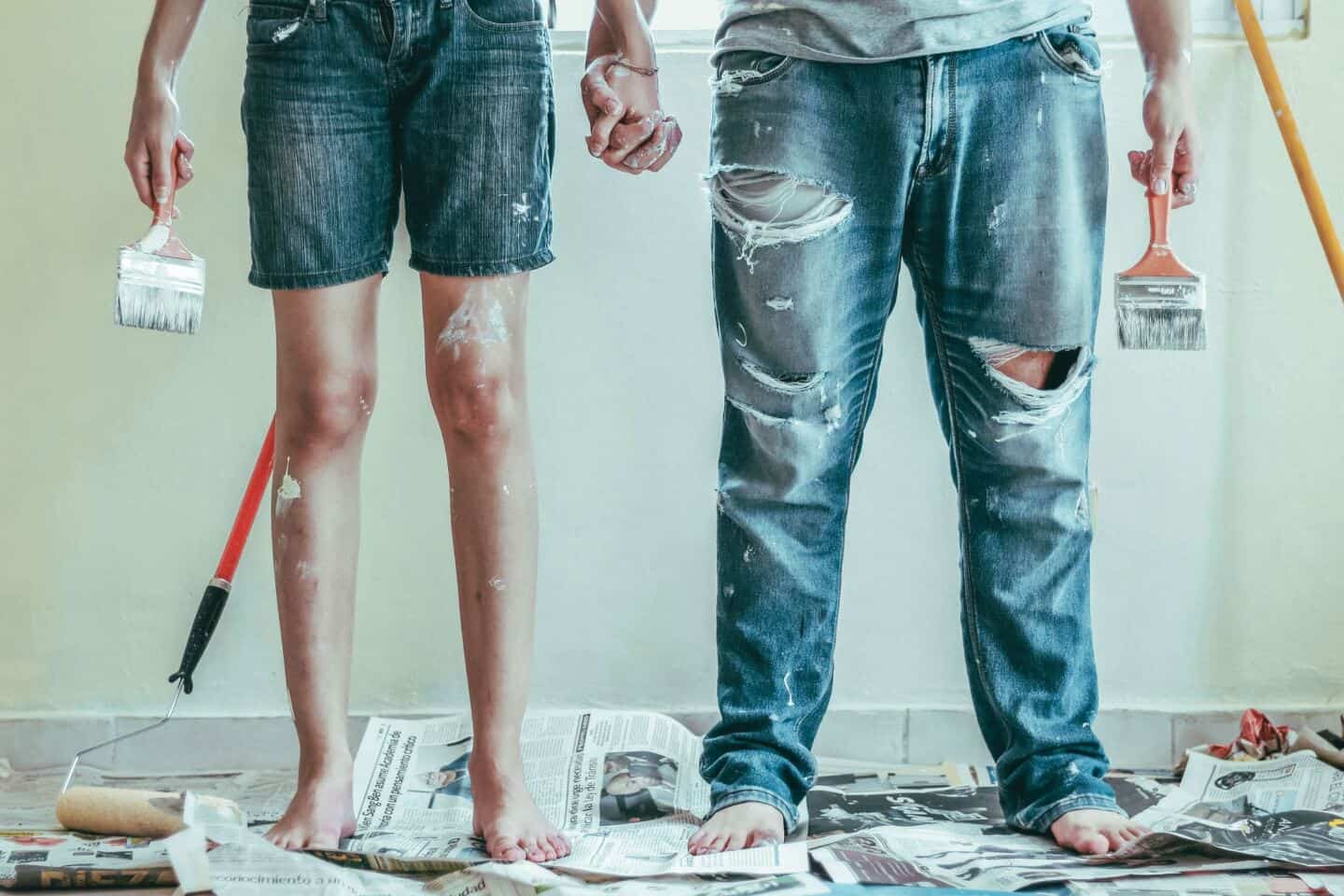 A young man and woman stand barefoot on newspaper holding hands and paint brushes ready to embark on a DIY home decor project